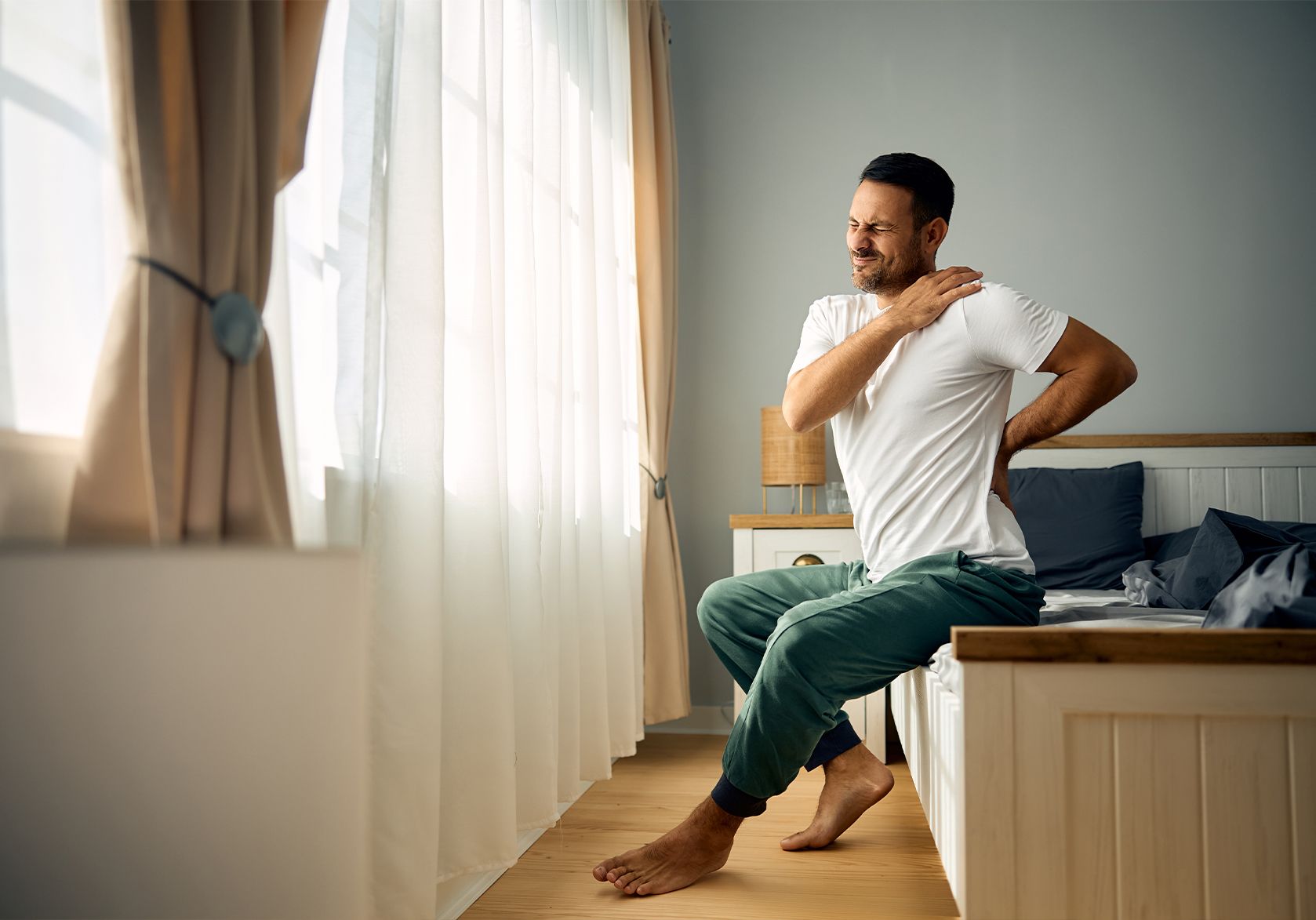 Hombre sentado en el borde de la cama tocándose el hombro con gesto de dolor. Representa cómo las lesiones articulares limitan las actividades cotidianas.